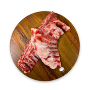 Two raw goat rib sections for raw feeding to dogs with visible bone and marbling, arranged on a round wooden cutting board against a plain white background.
